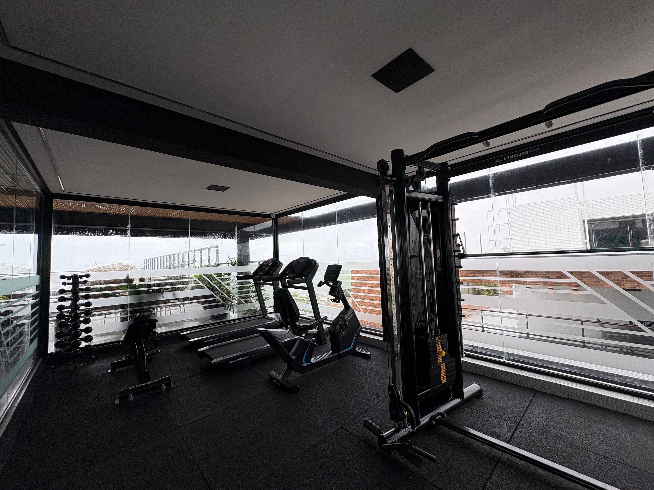 Ein Fitnessstudio mit Laufbändern, Kurzhanteln und Kraftgeräten, großen Fenstern und Blick auf ein Gebäude.