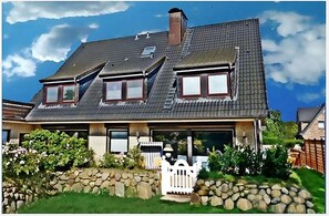 A two-story house with a sloped roof, a stone wall, and a white picket fence. - 4-Room Vacation Apartment "SYLTER DEICHWIESE" with Garden/Terrace in Westerland, Sylt (Sylt-Westerland)