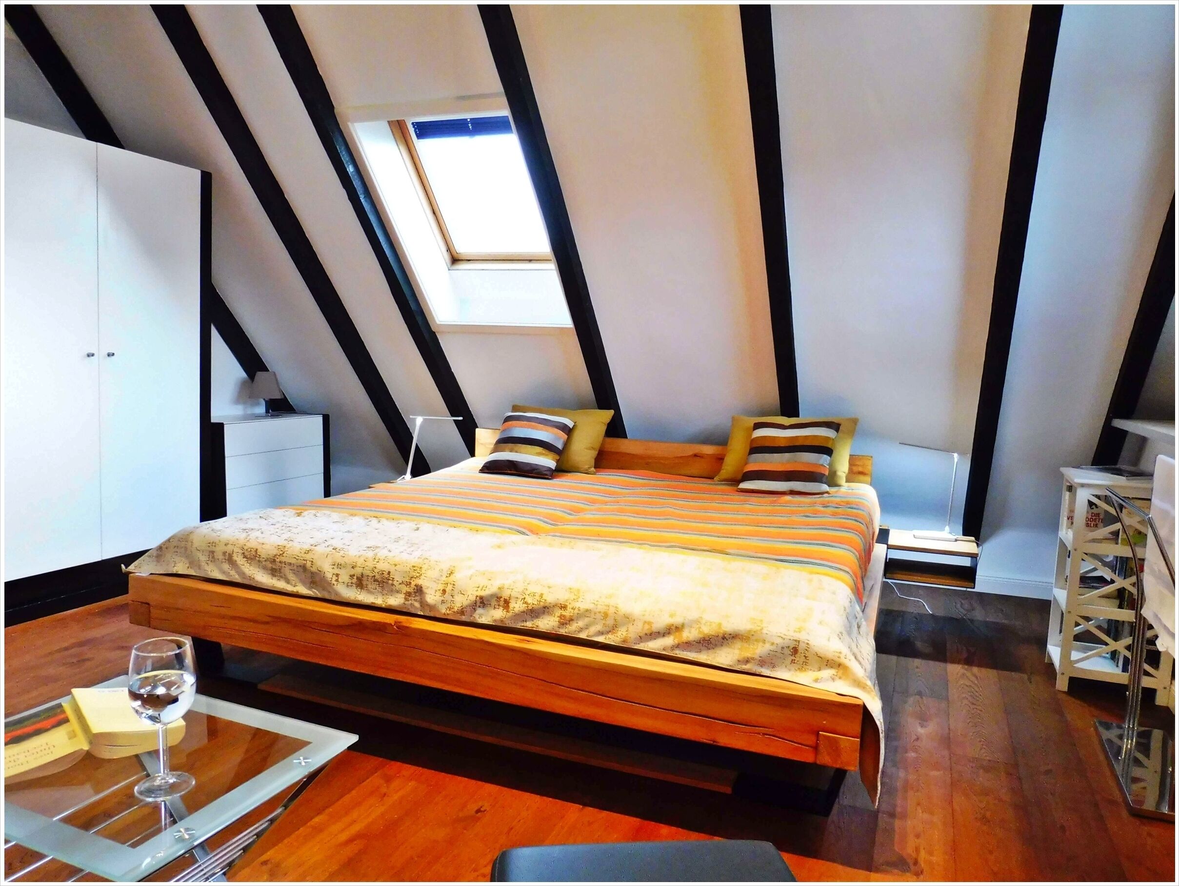 A bedroom with a sloped ceiling, a bed with striped bedding, a bedside table with a glass of wine, and a small side table with a lamp.