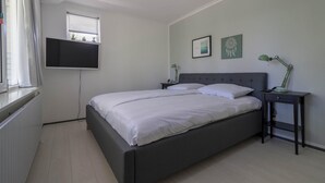 Un dormitorio con cama, mesita de noche, lámpara, televisor montado en la pared y ventana con persianas.