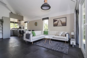 Ein modernes Wohnzimmer mit einem weißen Sofa, einem runden Couchtisch und einem grauen Teppich. Im Hintergrund befindet sich eine Küche mit Essbereich.