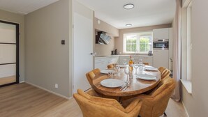 A dining area with a round wooden table and four chairs, a kitchen in the background, and a window with blinds. - Beach Villa With Garden & Water Access (Makkum)