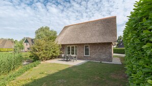 A house with a thatched roof, a small patio with a table and chairs, and a well-maintained lawn. - Beach Villa With Garden & Water Access (Makkum)