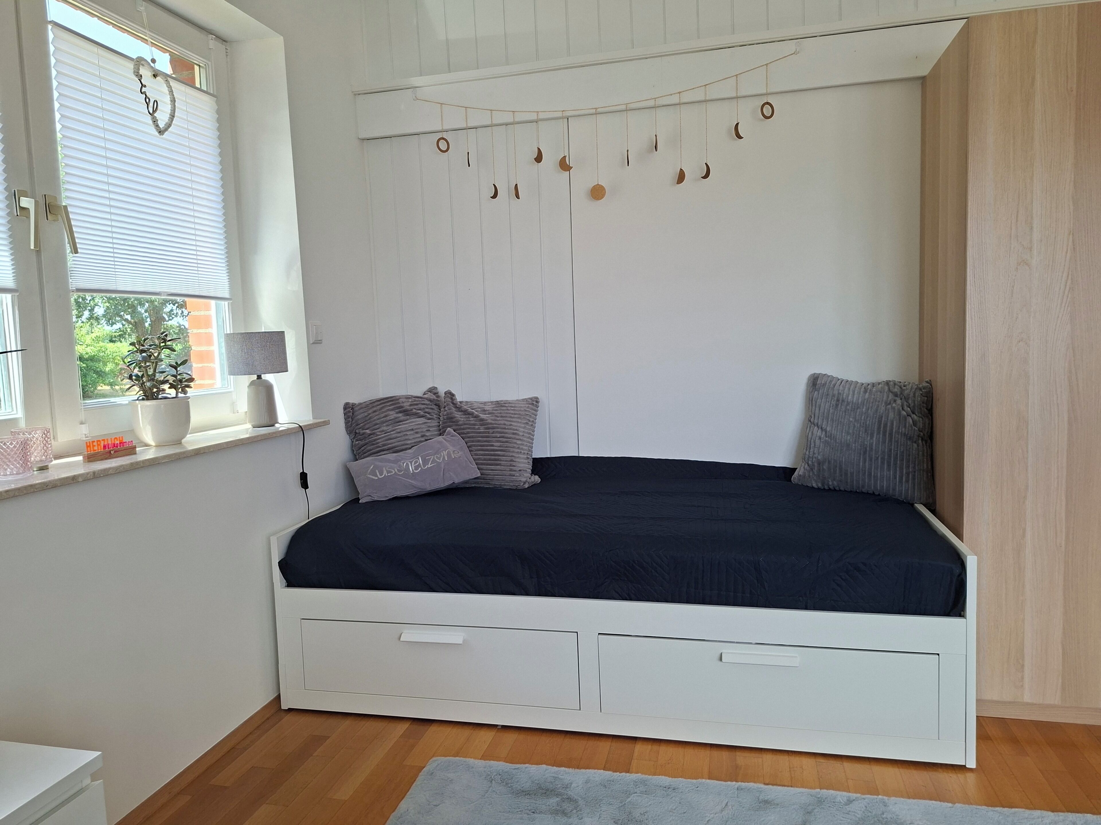 A bedroom with a bed, a window with blinds, a bedside table with a lamp, and a wall-mounted decoration.