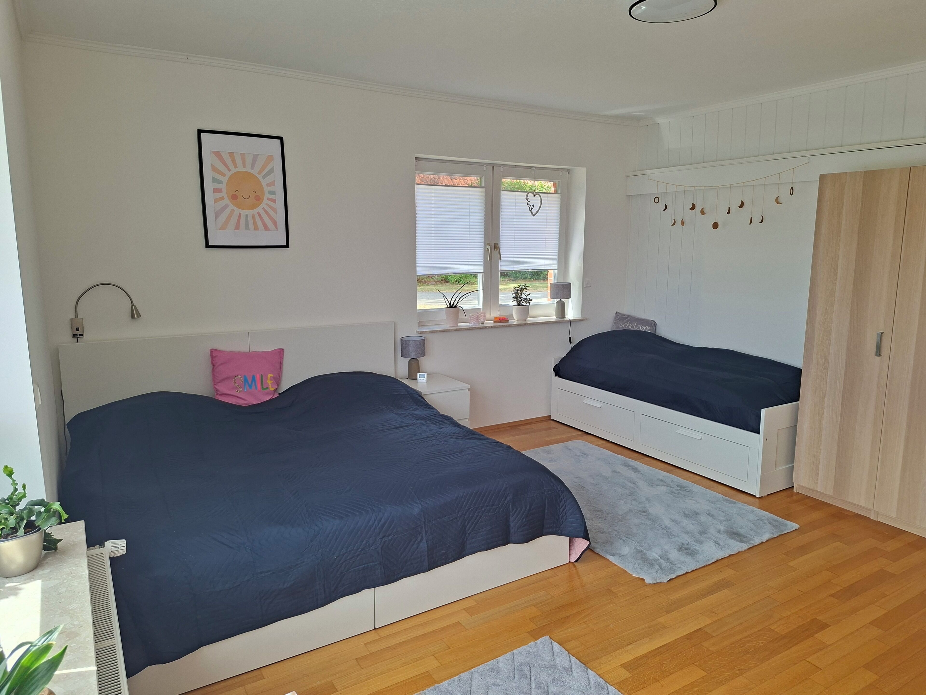 Ein Schlafzimmer mit zwei Betten, einem Fenster mit Jalousien, einem Nachttisch, einer Lampe, einem Heizkörper, einer Topfpflanze und einem Kleiderschrank.