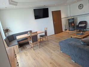 A modern living room with a wooden dining table, two chairs, a grey sofa, a television mounted on the wall, and a fireplace.