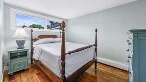 Un dormitorio con cama con dosel, una mesita de noche con lámpara, cómoda y ventana con vistas.