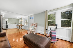Un salon spacieux doté d’un parquet, d’un canapé en cuir marron, d’un fauteuil à motifs floraux et d’un coin repas avec une table et des chaises en bois.