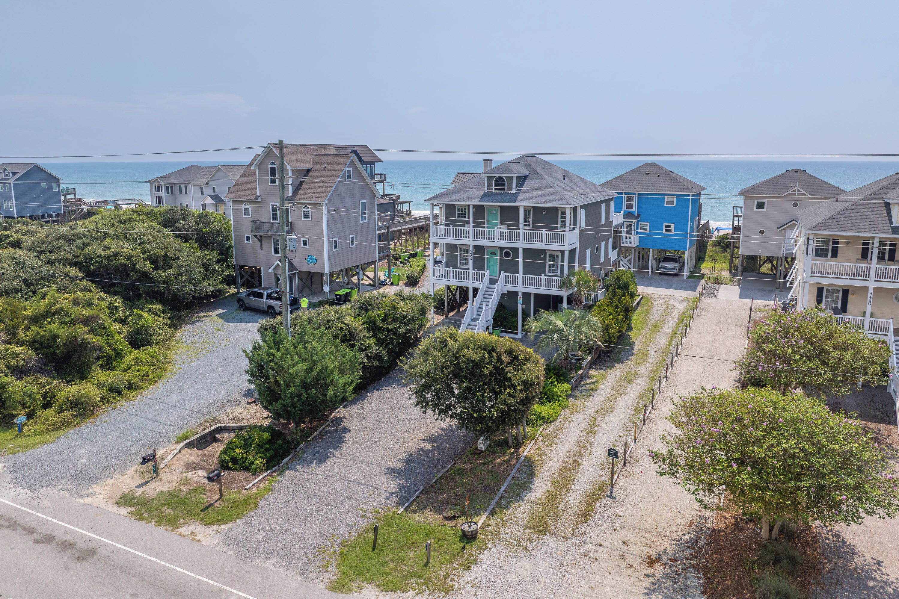 A coastal residential area with multi-story houses, a gravel driveway, and a clear view of the ocean.
