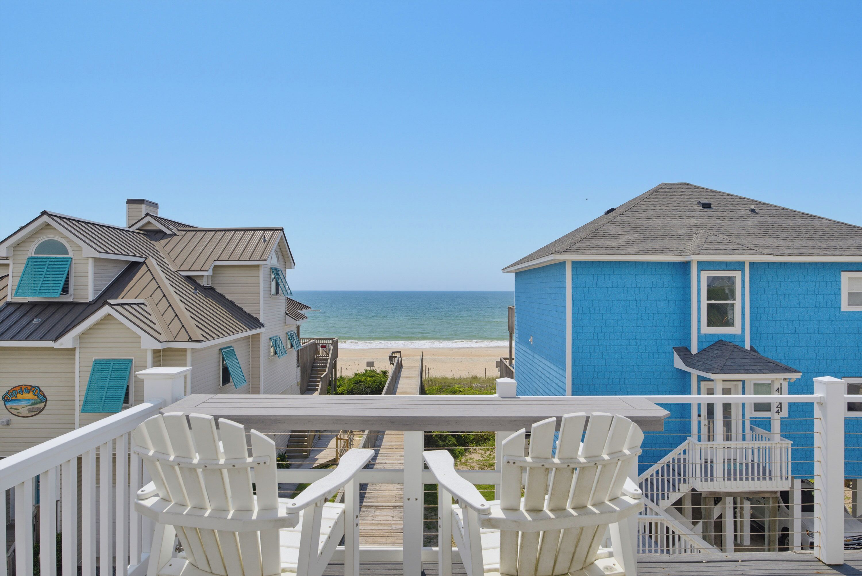 A wooden deck with white chairs, a view of the beach, and a blue building with a sign.