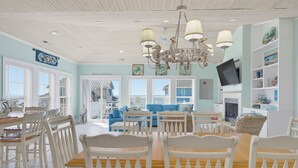 Et spiseområde med hvide træstole og et bord, en stue med blå sofaer og et fjernsyn, samt et loft med en unik lysekrans.