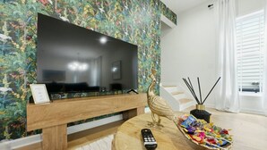 Ein Wohnzimmer mit Flachbildfernseher auf Holzfuß, einer dekorativen Pfauenstatue, einer Fernbedienung und einer Schale mit bunten Süßigkeiten auf einem runden Holztisch.