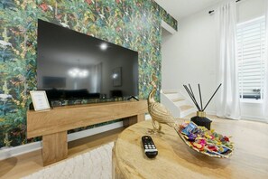A living room with a flat-screen TV on a wooden stand, a decorative peacock statue, a remote control, and a bowl of assorted candies on a round wooden table. - West End Gem | Near Mercedes-Benz Stadium, Pet OK (Atlanta)