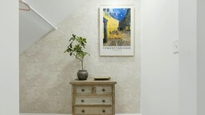 Ein Zimmer mit strukturierter Wand, einer Holzkommode mit Schubladen, einer Topfpflanze und einem gerahmten Kunstwerk, das eine Straßenszene zeigt.