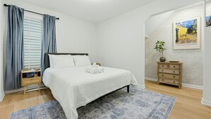Ein Schlafzimmer mit Bett, Nachttisch, Kommode, einem Fenster mit Vorhängen und einem Wandbild.