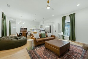 Un salon moderne avec un canapé, un pouf et un fauteuil en forme de sac. On y trouve également un coin repas avec des chaises et une table. Au fond de la pièce, il y a une cuisine avec des meubles blancs et un réfrigérateur.