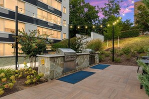 An outdoor patio with a stone barbecue and grill, surrounded by a landscaped garden and a building in the background. - Gracious 2 Bed 2 Bath Apartment with Free Parking (Arlington)