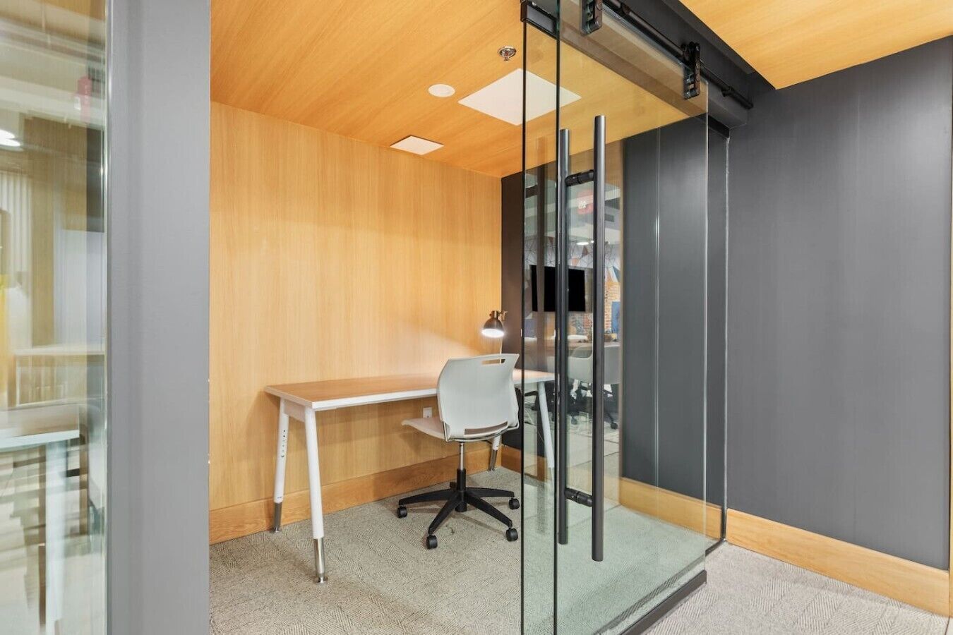 Un espacio de oficina moderno con un escritorio y una silla de madera, un separador de vidrio y una pared gris.