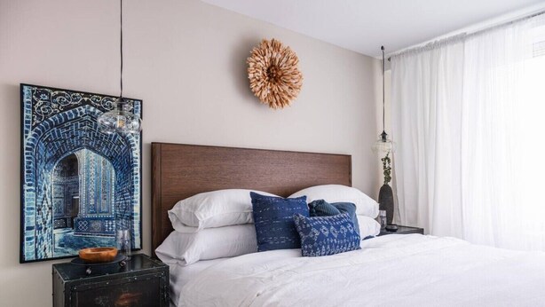 A bedroom with a large bed, a headboard, a wall-mounted decorative piece, a nightstand with a lamp, and a framed artwork. - Gracious 2 Bed 2 Bath Apartment with Free Parking (Arlington)