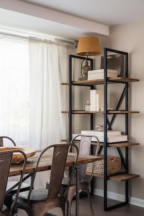 A dining area with a wooden table and chairs, a metal bookshelf, and a window with sheer curtains. - Gracious 2 Bed 2 Bath Apartment with Free Parking (Arlington)