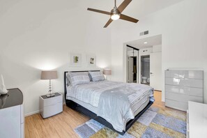 A bedroom with a bed, nightstands, a dresser, a ceiling fan, and a door leading to a bathroom. - Rose Garden Escape - Stylish 2 Bedroom Condo (Palm Springs)