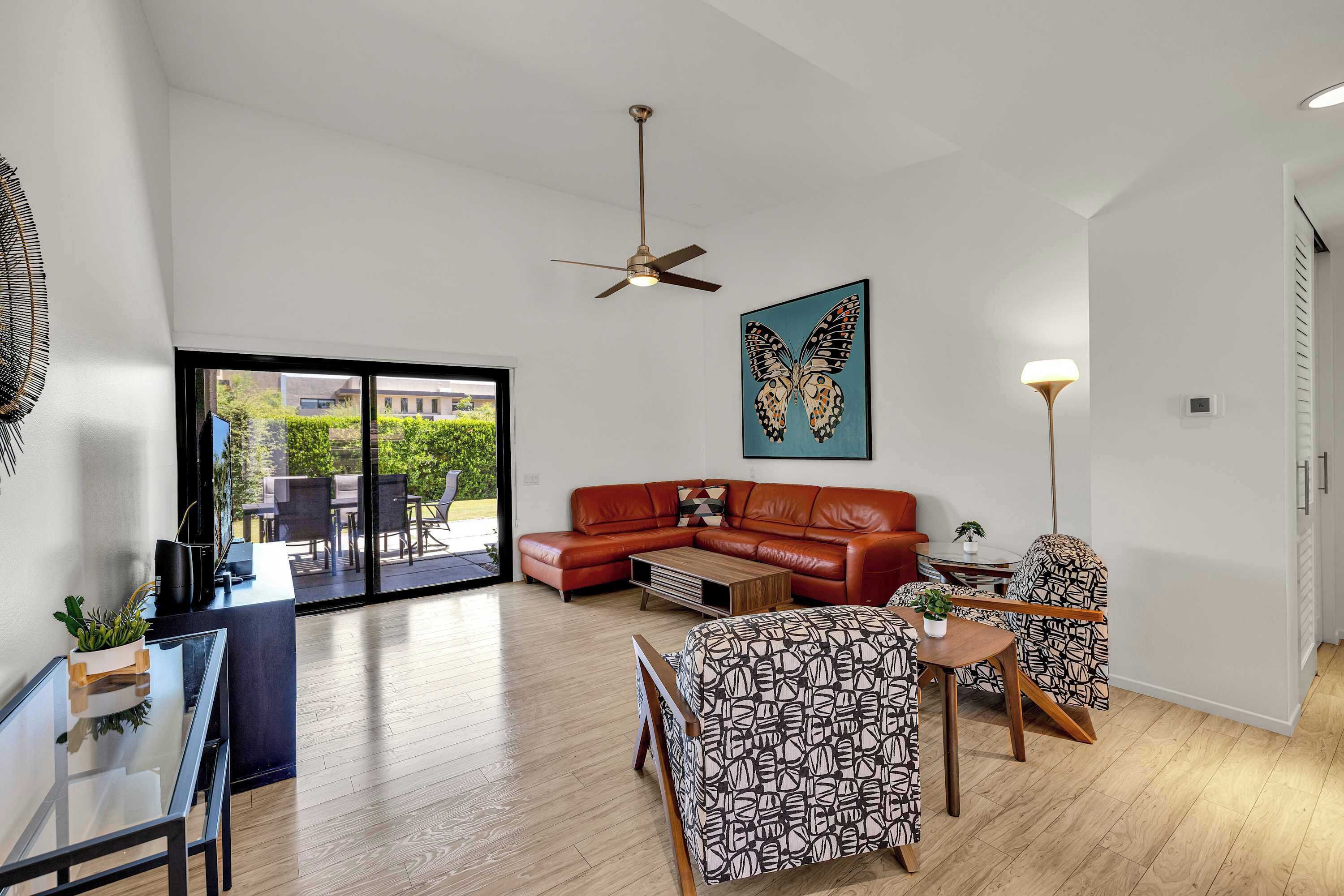Una sala moderna con sofá de cuero, una mesa de centro, sillas estampadas, ventilador de techo y un amplio ventanal con vistas al exterior.