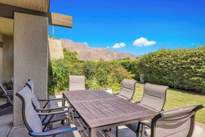 An outdoor patio with a wooden table and chairs, surrounded by greenery and a mountain view. - Rose Garden Escape - Stylish 2 Bedroom Condo (Palm Springs)