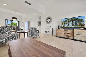 Un salon moderne avec une table à manger en bois, des canapés à motifs, un téléviseur et une vue sur des palmiers.