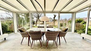 Et orangeri med glasfacade, et rundt bord, fletstole og en lampe.