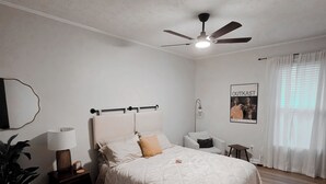 Ein Schlafzimmer mit einem Bett, Nachttischen, einem Deckenventilator, einem Poster an der Wand und einem Fenster mit Vorhängen.