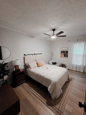 Une chambre à coucher avec un lit, des tables de chevet, un ventilateur de plafond, une affiche au mur et une fenêtre avec des rideaux.