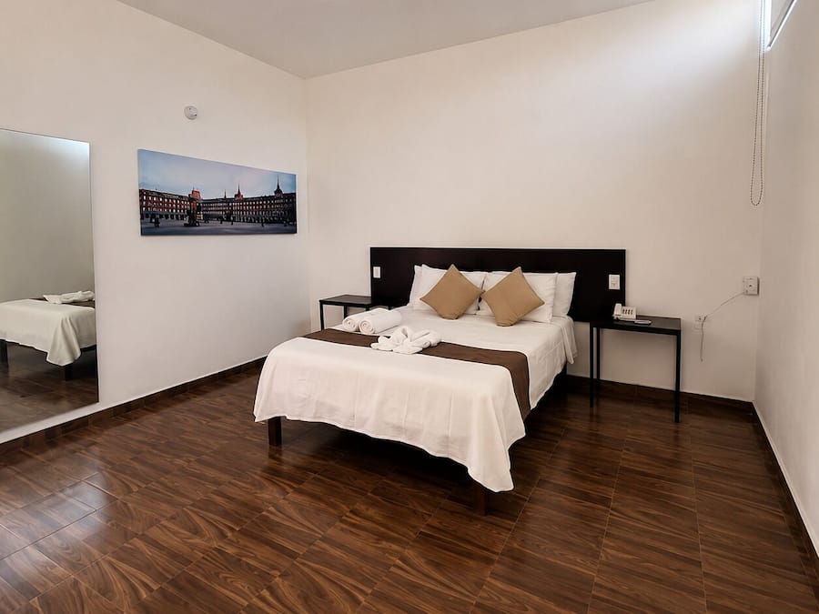Una habitación de hotel con cama, mesitas de noche, un espejo y un cuadro colgado en la pared.