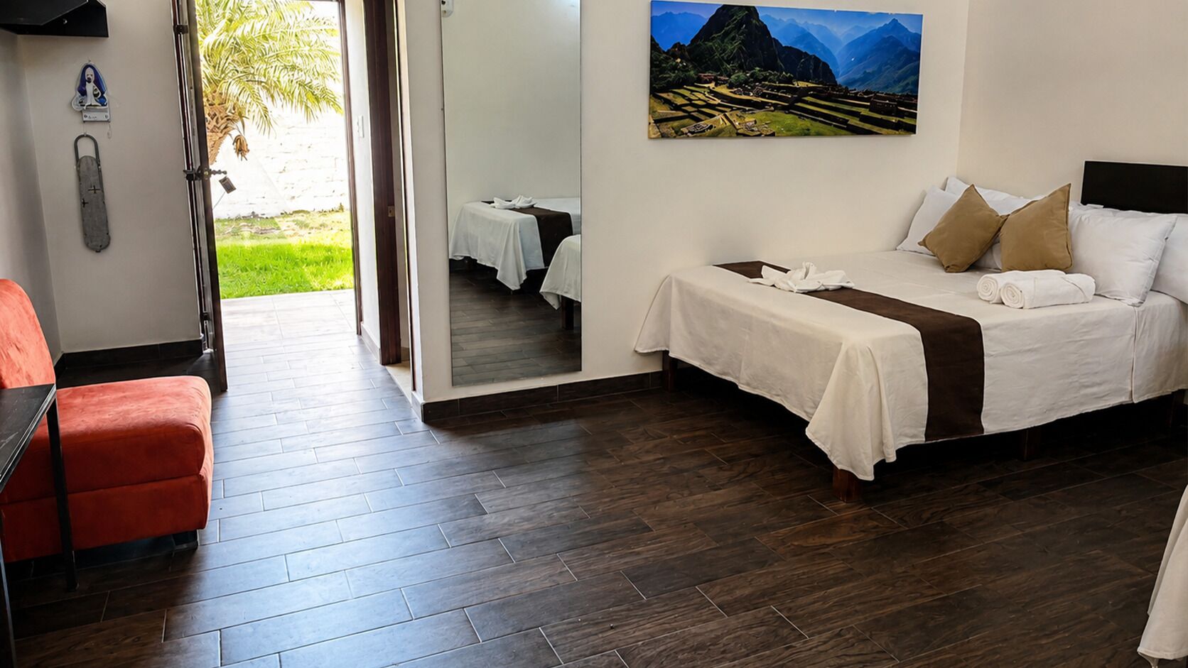 A hotel room with a bed, a red chair, a wooden floor, and a painting of a mountainous landscape.