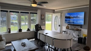 A modern living room with a dining area, large windows, a ceiling fan, and a flat-screen TV.