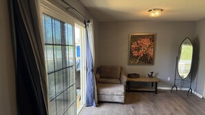 A room with a sliding glass door, a brown leather sofa, a small wooden table, a decorative mirror, and a framed floral painting on the wall.