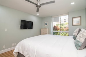 Ein Schlafzimmer mit Bett, Deckenventilator, Wandfernseher und Fenster mit Blick auf Gebäude und Grünflächen.