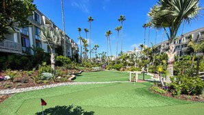 A golf course with a putting green, surrounded by palm trees and a building with balconies. - Come Stay at This Coastal Comfort in the Heart of Socal (Oceanside)