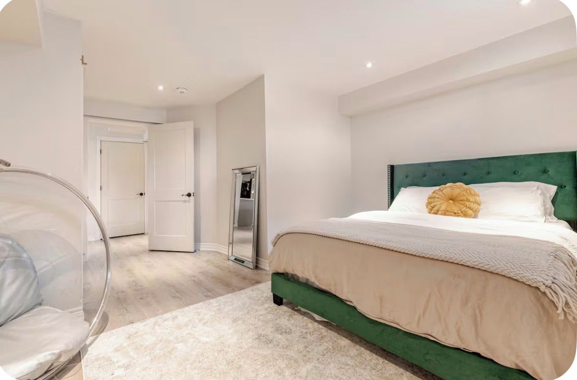 A bedroom with a green upholstered headboard, a large mirror, and a clear plastic chair.