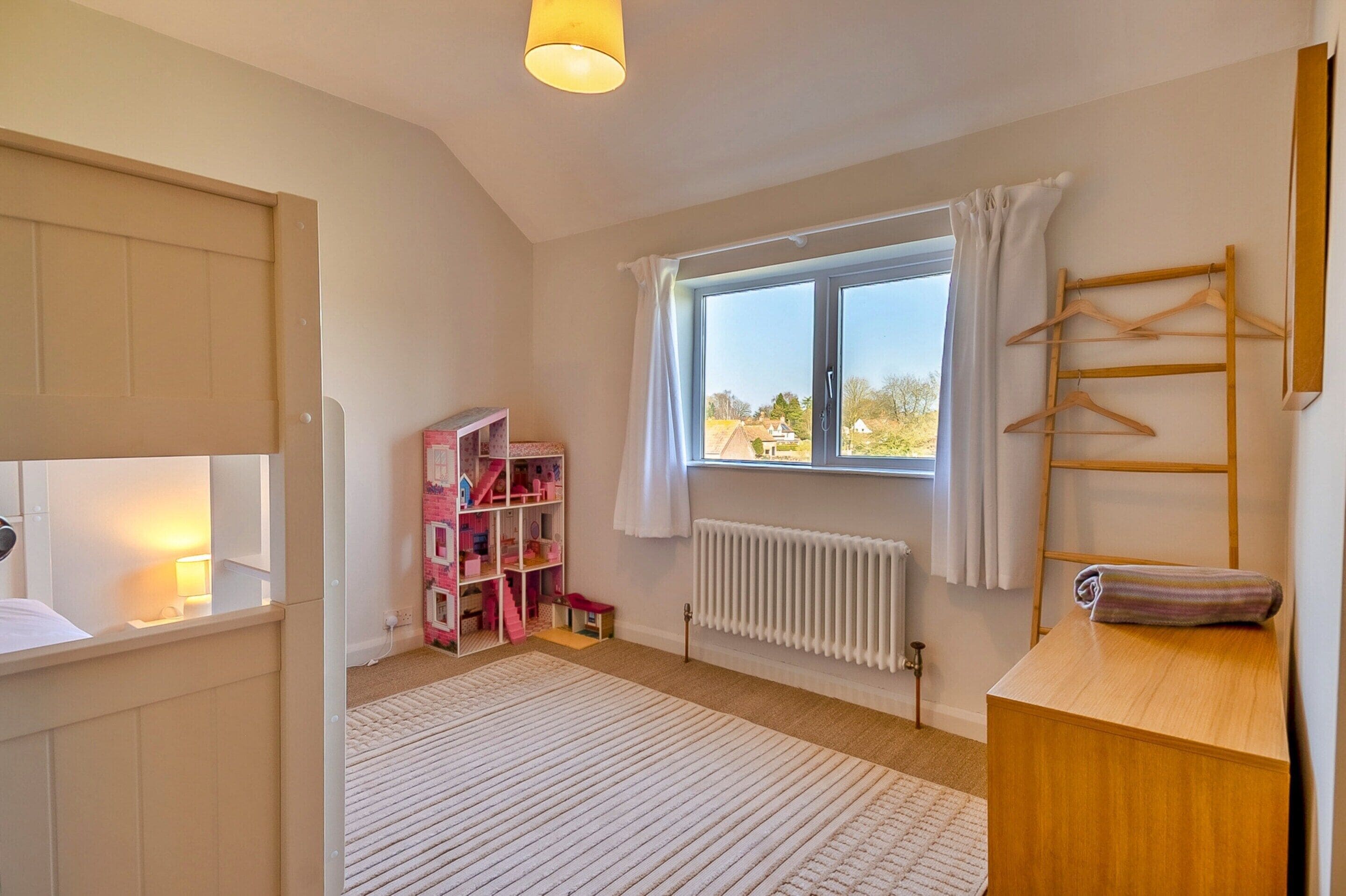 Una habitación con una cama, un radiador, una ventana con cortinas, un aparador de madera y una casa de juguete rosa.
