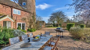 A dining area with a table set for four, outdoor seating, a barbecue grill, and a brick building with a tower in the background. - Unique Norfolk Windmill Retreat for 8 (Dereham)