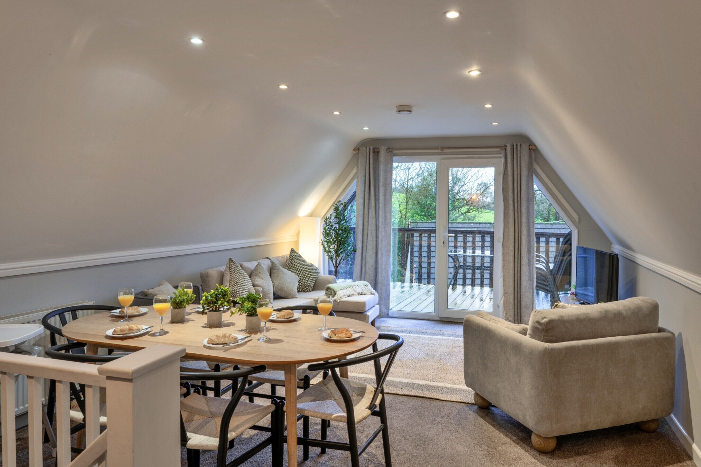 A dining area with a round table, chairs, and a sofa. There is a view of a deck through a glass door.