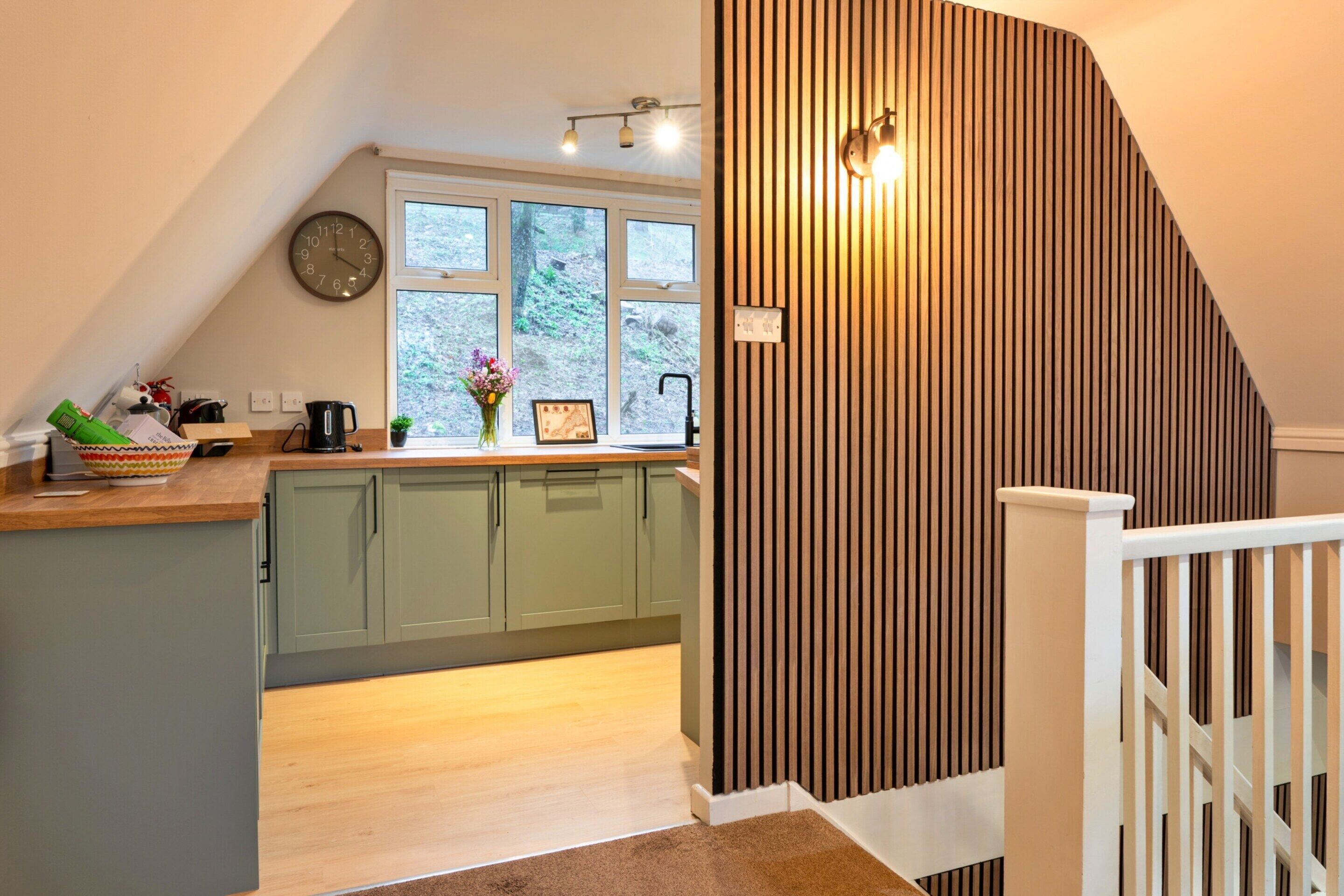 A kitchen with a wooden countertop, green cabinets, a clock, and a window with a view of trees.