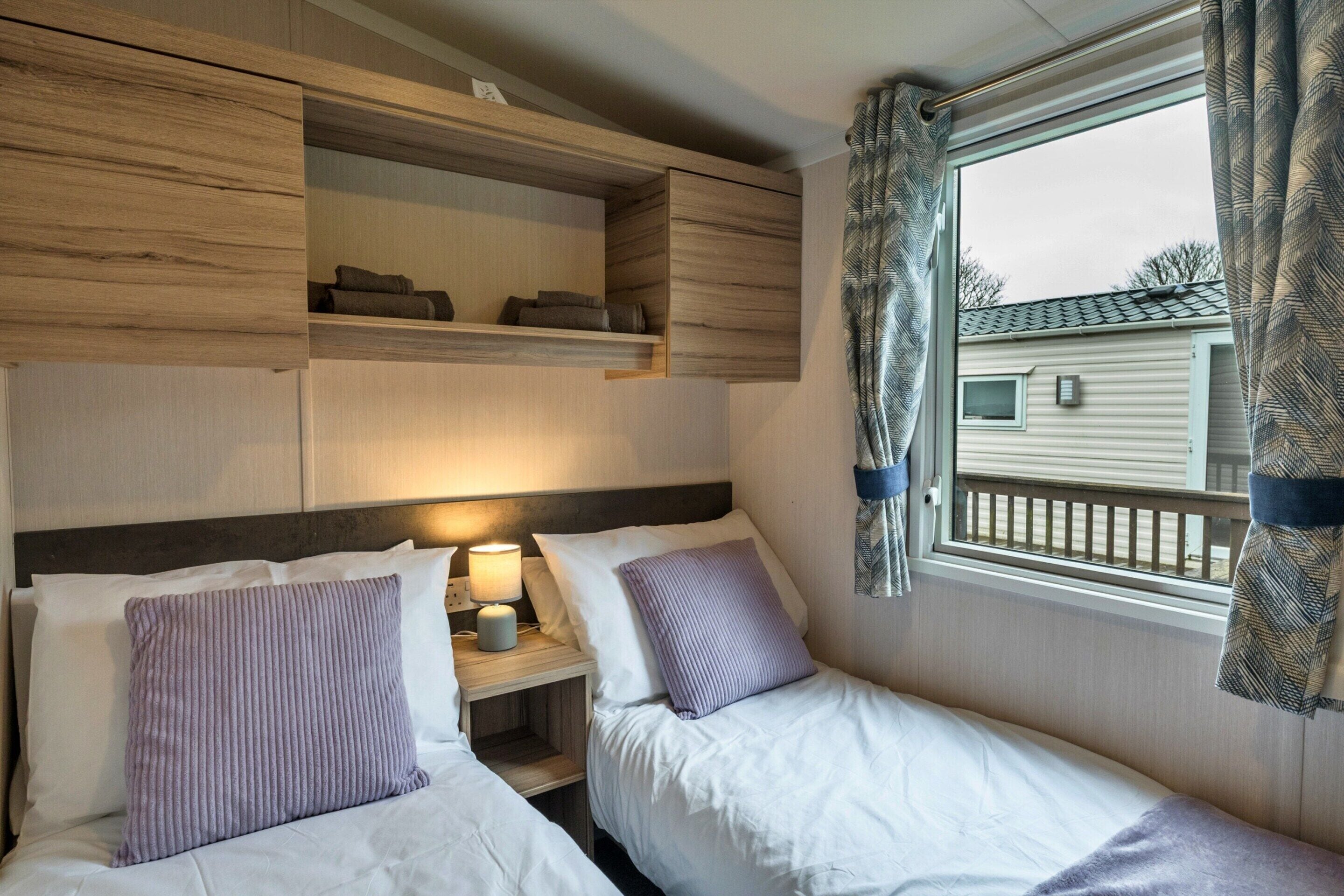 A bedroom with two beds, a nightstand, a window with curtains, and a built-in shelf above the beds.