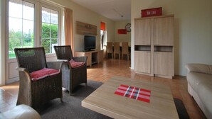 Un salon avec des meubles en osier, un téléviseur, un coin repas avec des chaises et un coin cuisine avec des placards.