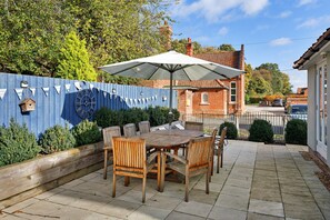 A patio with a wooden table and chairs, a blue fence with a clock, and a brick house in the background. - Charming Chapel House Norfolk (Dereham)