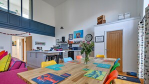 Eine moderne Küche mit einer zentralen Kochinsel, ein Essbereich mit bunten Untersetzern und ein Wohnbereich mit Sofa und Kissen.