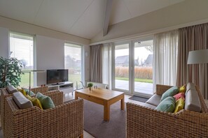 A spacious living room with a television, wicker furniture, a wooden coffee table, and large windows with curtains. - Elegant Waterfront Bungalow With Jetty (Makkum)
