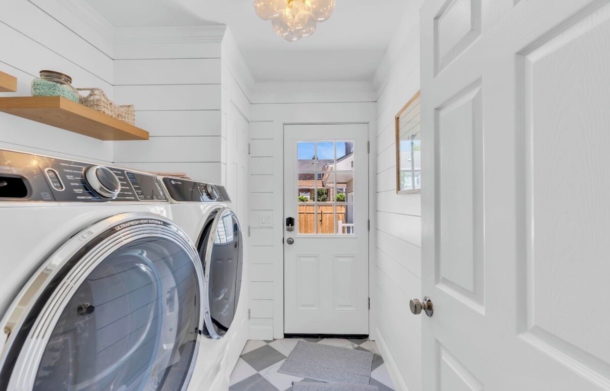 A laundry room with two front-loading washing machines, a wooden shelf with jars, and a white door leading to an outdoor area.