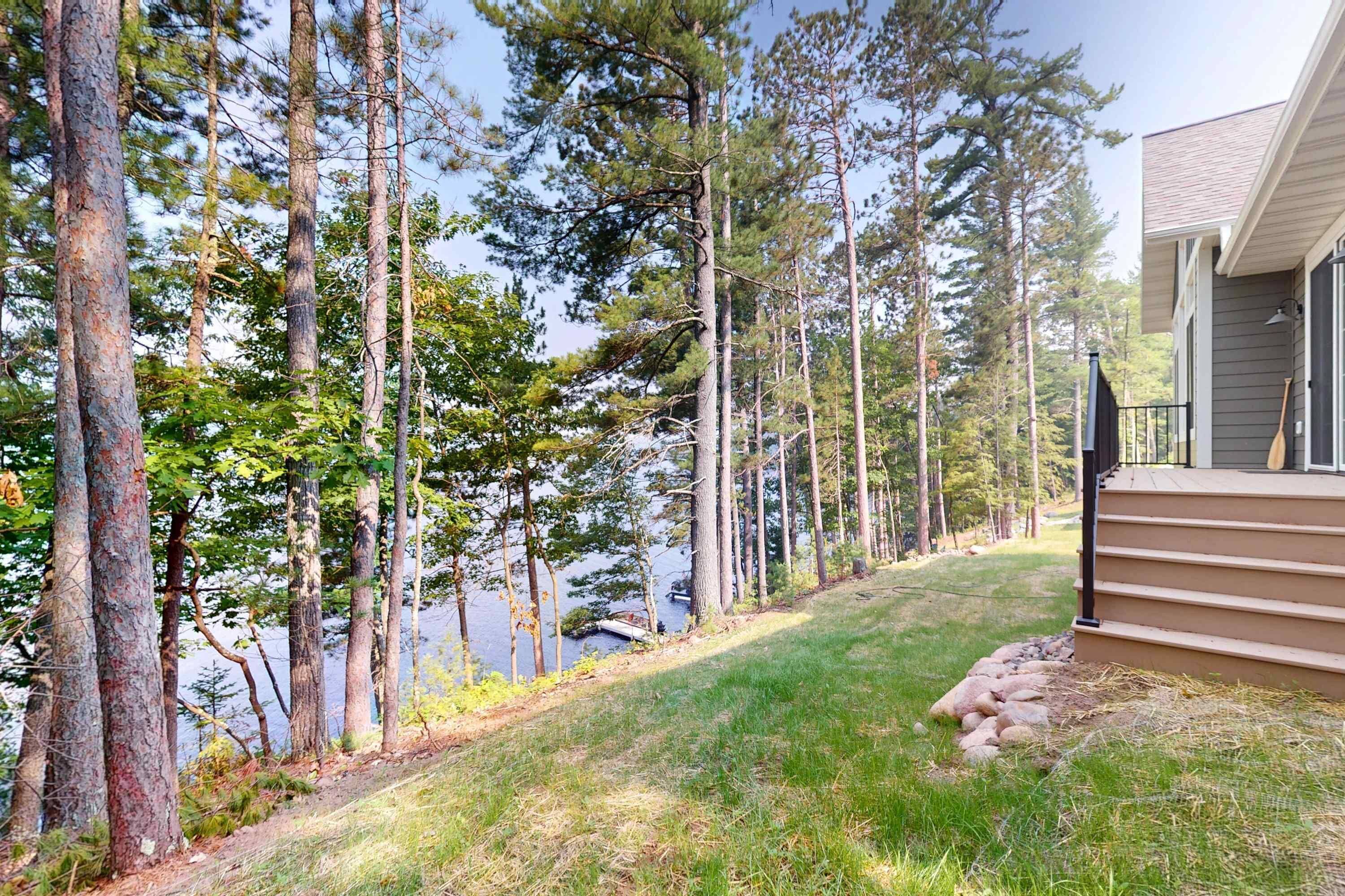 A residential property with a grassy yard, tall pine trees, and a dock extending into a body of water.