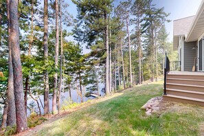 A residential property with a grassy yard, tall pine trees, and a dock extending into a body of water.